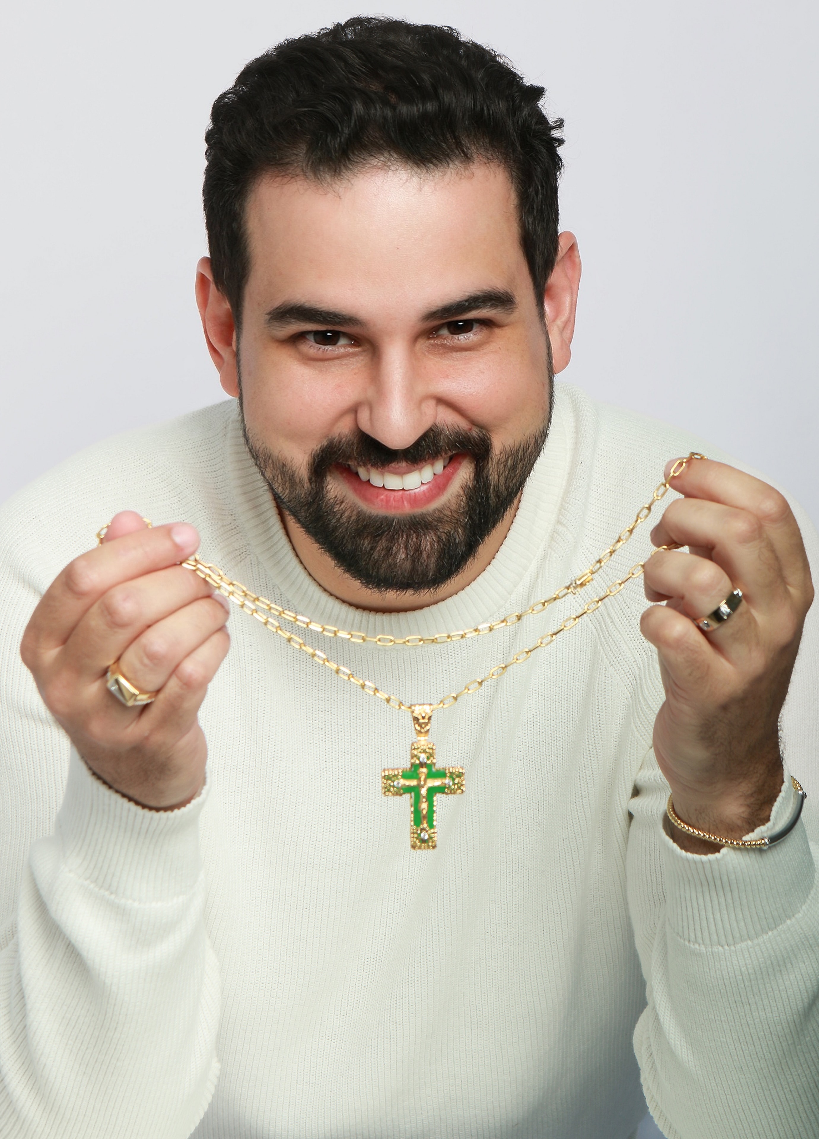 Danillo wearing a white sweater holding a gold necklace with green crucifix and rings